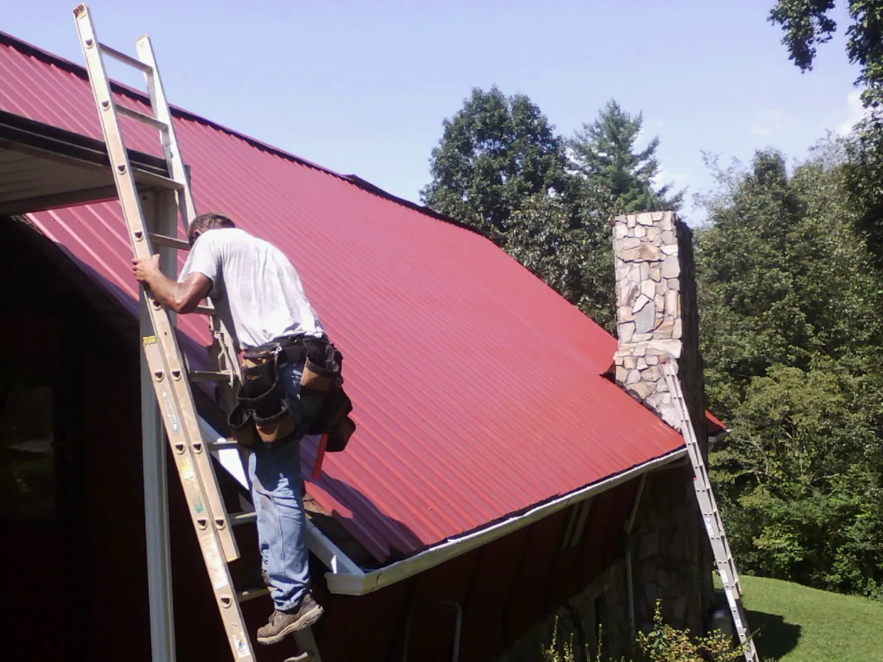 Roofer on a ladder beside a red metal roof and stone chimney — Soffit & Fascia Repair by Roofing Brothers in Keenesburg